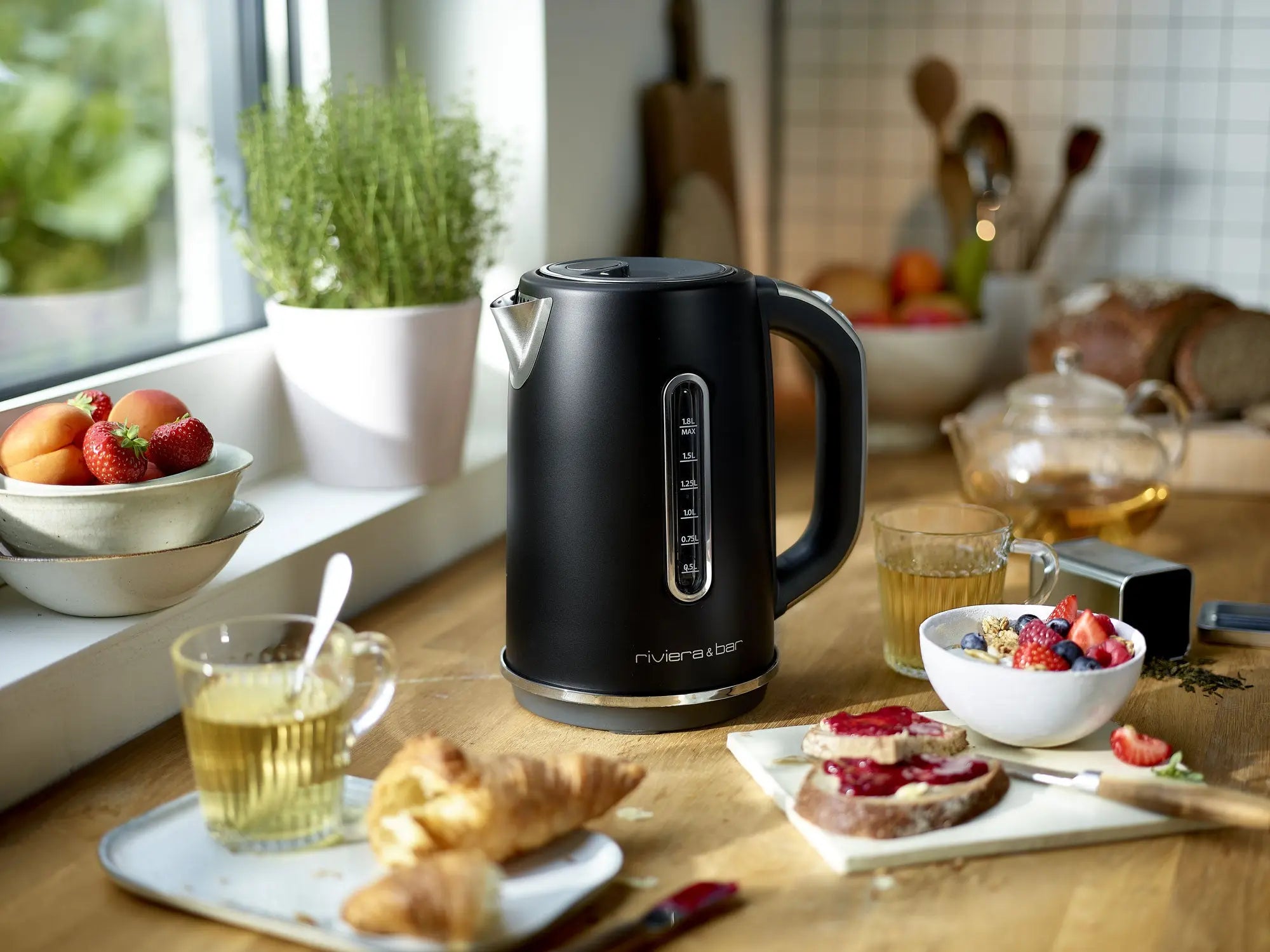 La Bouilloire Alma - Black Satin (1,8 L) by riviera&bar est posée sur un comptoir de cuisine en bois au milieu de thé, croissants, toasts à la confiture, baies, fruits et herbes près d'une fenêtre éclairée par le soleil, ajoutant de l'élégance à la scène du petit déjeuner.