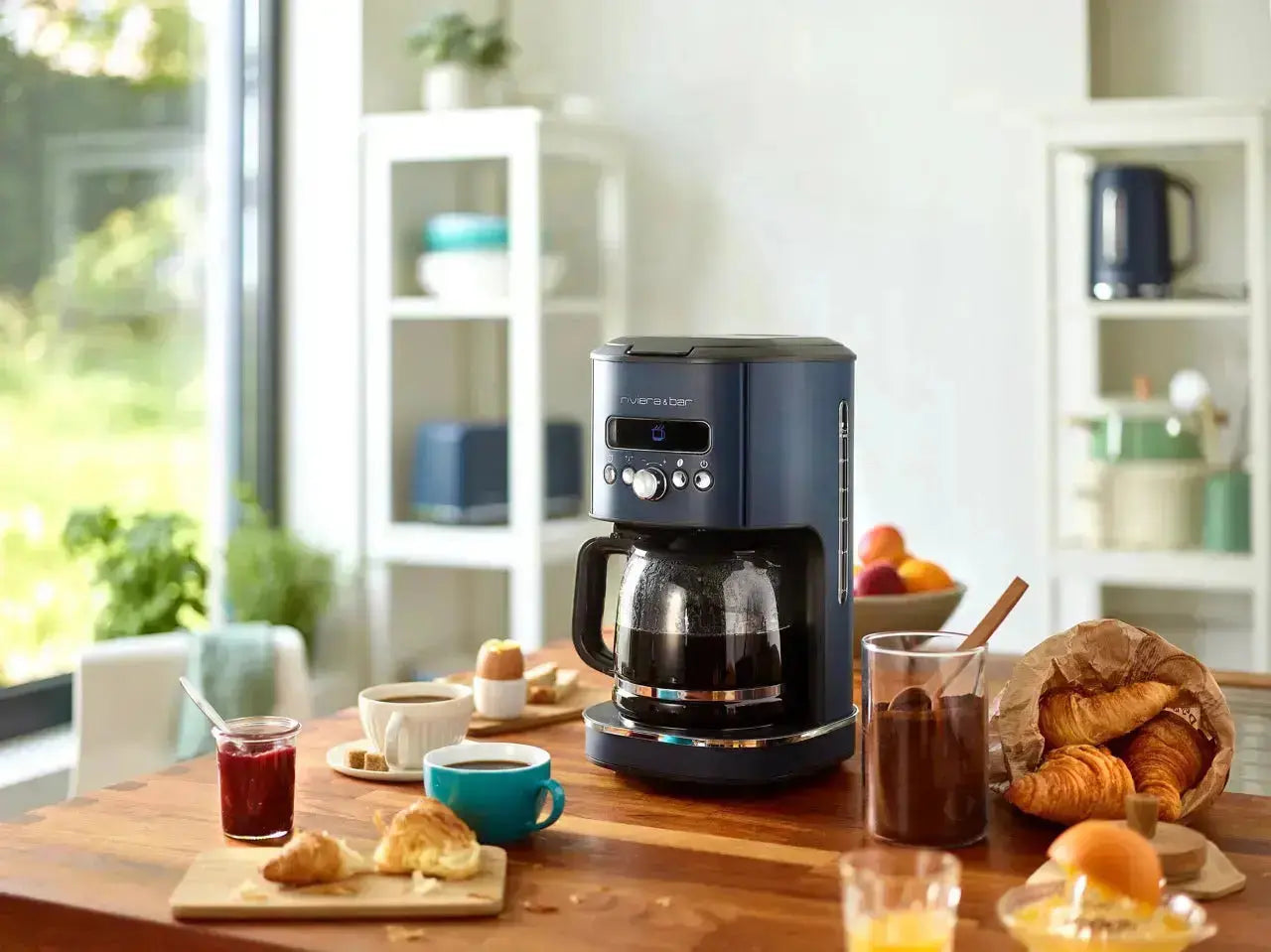 La riviera&bar Cafetière programmable BCF 578 - Bleu gris, 1,8 L est posée sur une table de cuisine en bois avec du jus d'orange, des croissants, de la confiture et des tasses à proximité. Des étagères et une fenêtre éclairent l'arrière-plan.