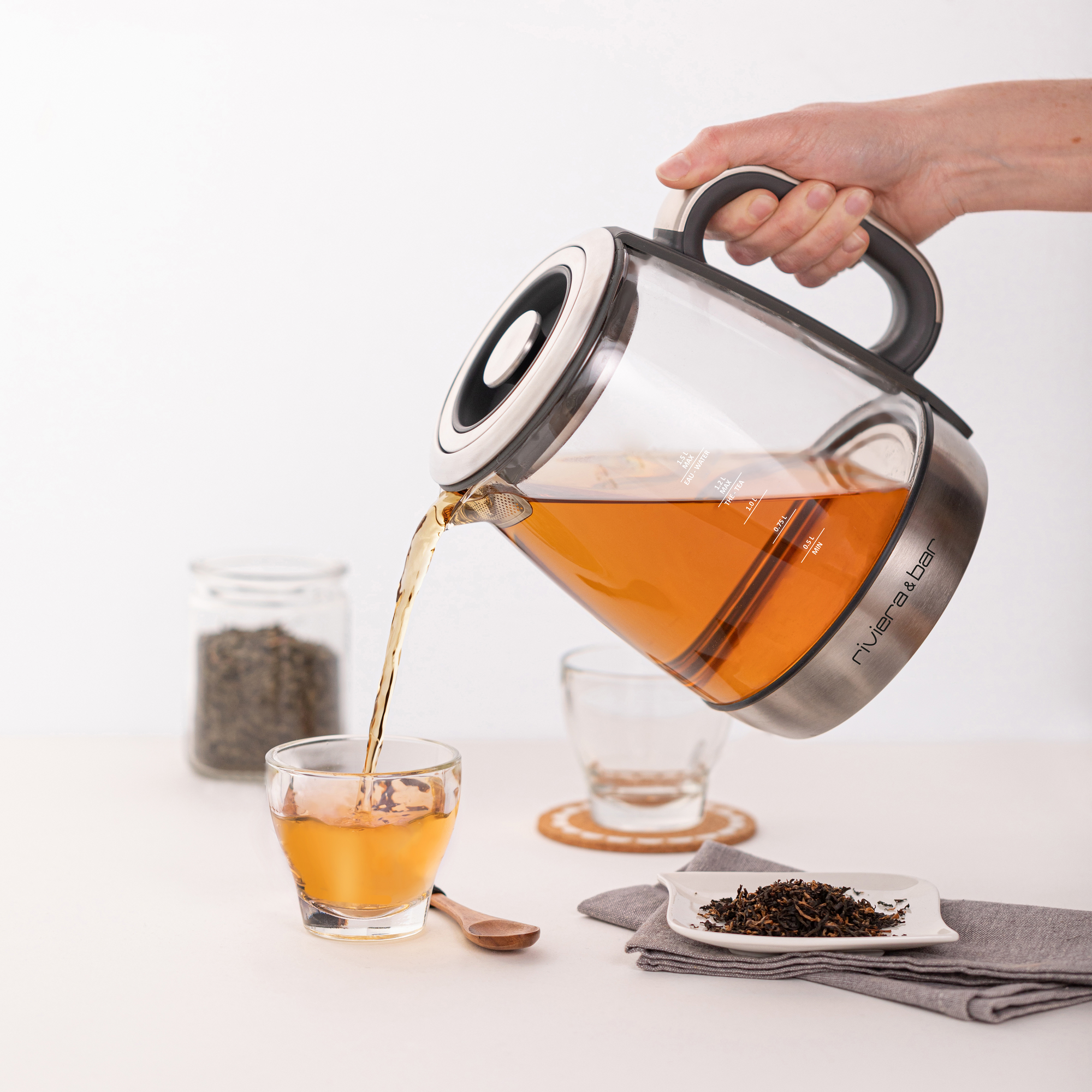 Une main verse du thé du riviera&bar Théière Cha Dao Expert dans une tasse sur une table, avec des feuilles de thé en vrac dans un pot et sur une assiette à proximité. Une cuillère, une tasse en verre vide et le panier à thé Monte et Baisse sont également visibles.