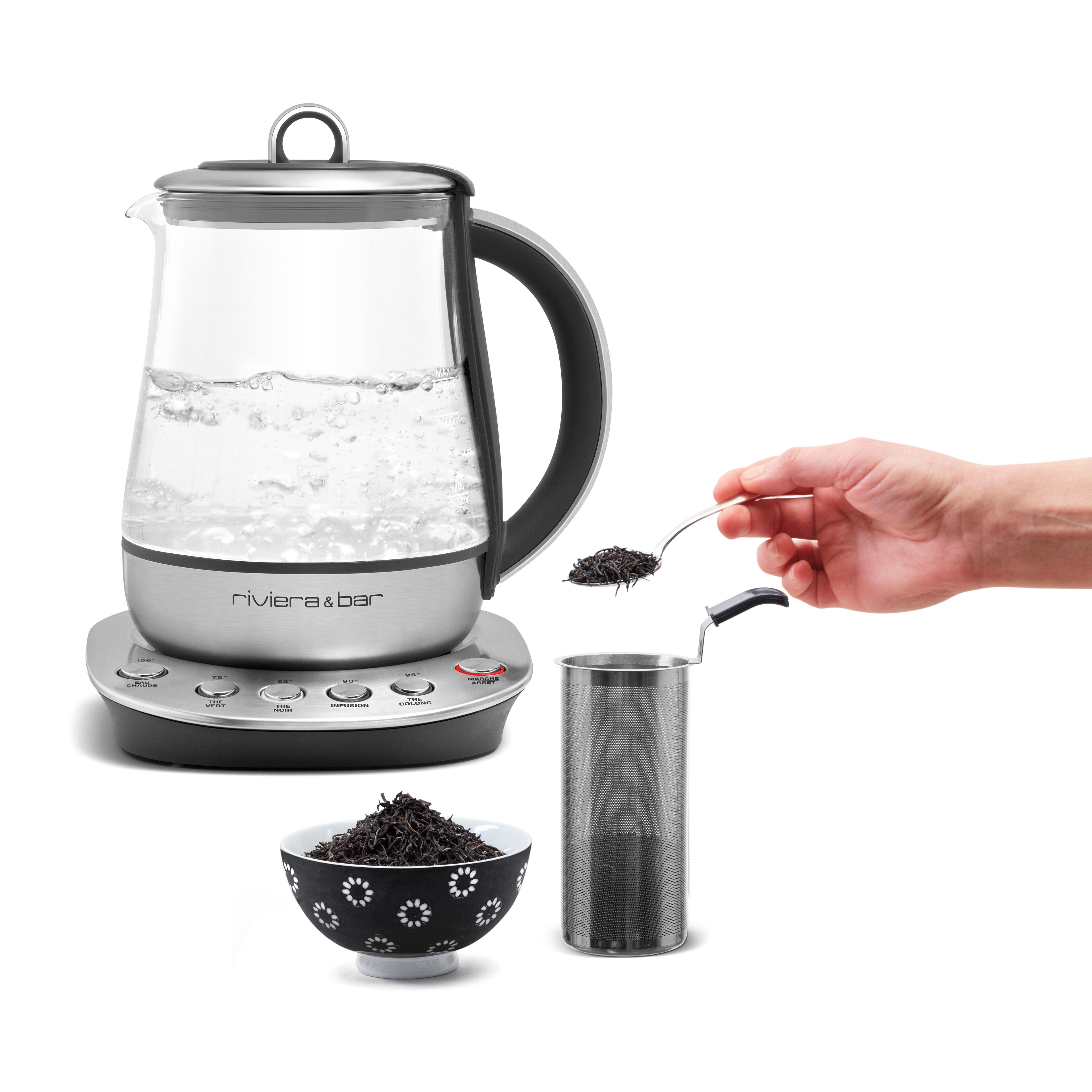 Une Théière Jaïpur Zen (1,2 L) de riviera&bar remplie d'eau bouillante se trouve à côté d'une main préparant du thé dans un infuseur en métal, tandis qu'un bol décoratif de feuilles de thé en vrac est présenté au premier plan.