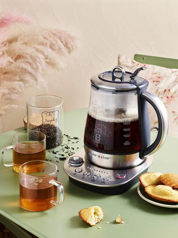 Image de présentation d'une théière électrique Riviera & Bar en verre et acier inoxydable, accompagnée de tasses de thé, d'une boîte de thé Darjeeling et de madeleines, sur une table verte.

