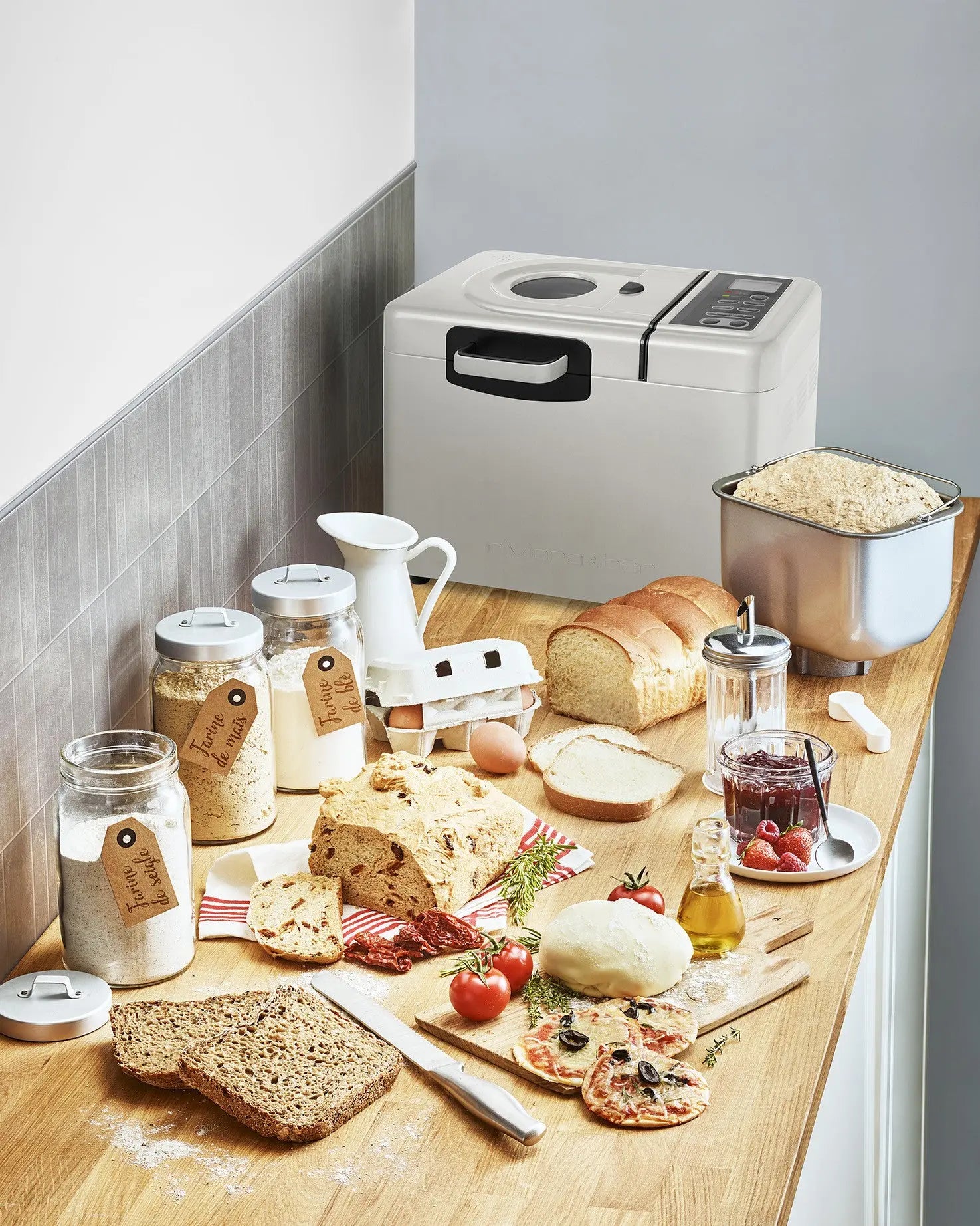 Une riviera&bar Machine à Pain Sans gluten & Co MP579A est posée sur un comptoir en bois, entourée de farine sans gluten, d'œufs, de lait, de tranches de pain fait maison, de pâte, de pots d'huile et de confiture, d'herbes, de tomates et de fromage - mettant en valeur ses résultats de cuisson.