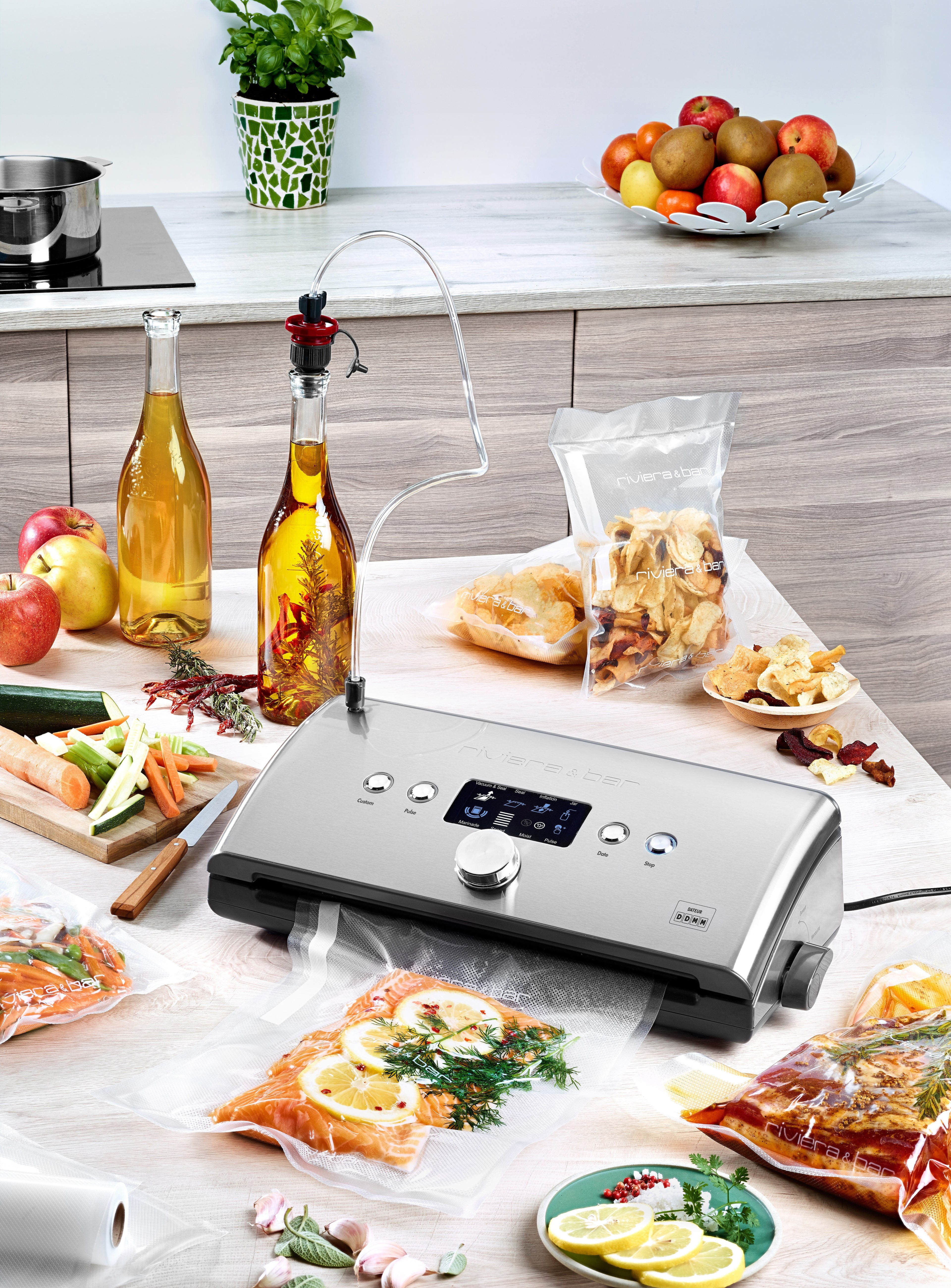 L'Appareil de mise sous vide avec dateur intégré riviera&bar emballe des sacs de saumon mariné aux herbes et citron sur un comptoir, entouré de fruits, légumes, épices et huiles pour conserver les aliments et limiter l'oxydation.
