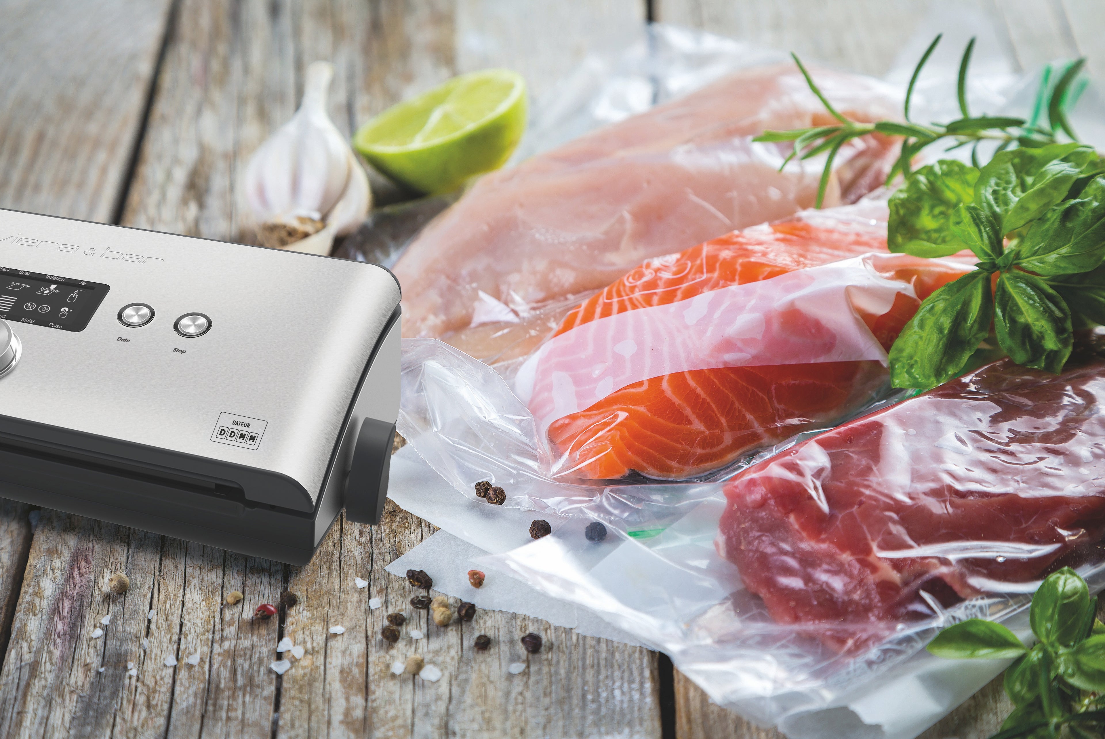 L'Appareil de mise sous vide avec dateur intégré riviera&bar est présenté en train de sceller un sac contenant poulet, saumon et bœuf, entourés d'herbes, ail, citron vert et poivre pour préserver les aliments et éviter l'oxydation.