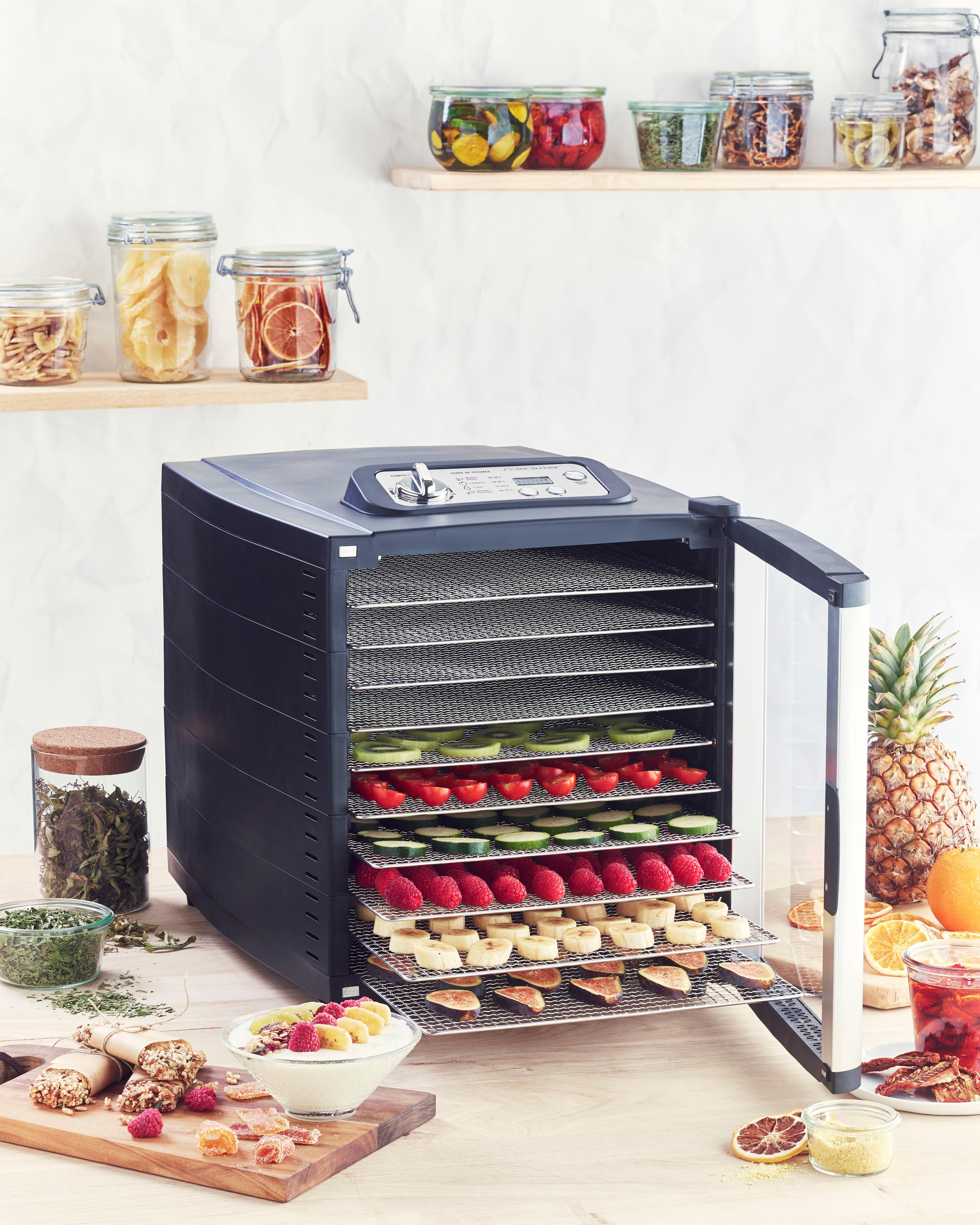 Le Déshydrateur alimentaire riviera&bar avec minuteur numérique et plateaux ajustables est ouvert, dévoilant fruits et légumes tranchés. Autour, bocaux de conserves et ingrédients frais sont disposés sur des étagères et une table.