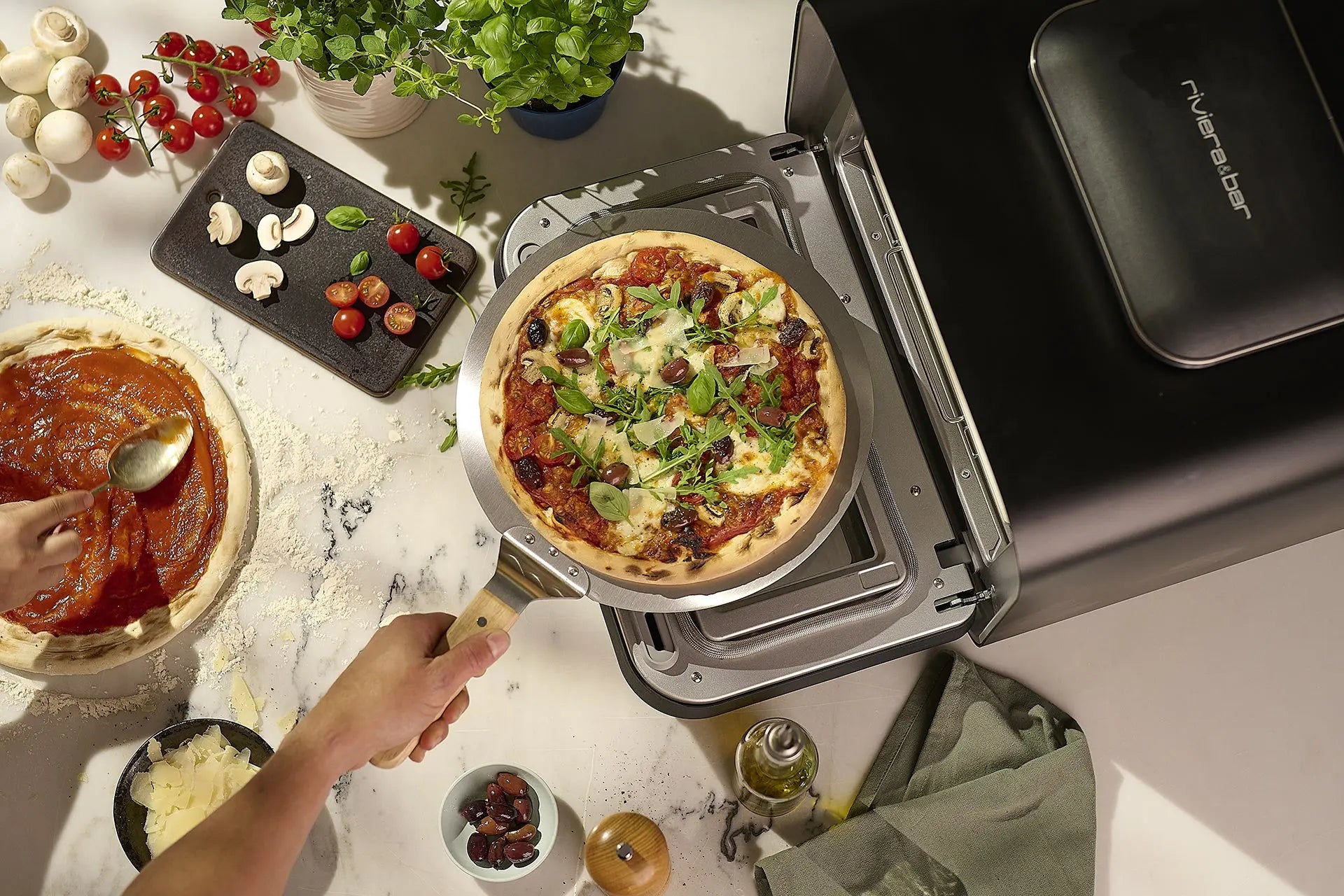 Quelqu'un glisse une pizza maison avec roquette et champignons dans un riviera&bar Four à pizza électrique 400 degrés, tandis que d'autres ingrédients et un autre plat italien sont préparés à proximité.