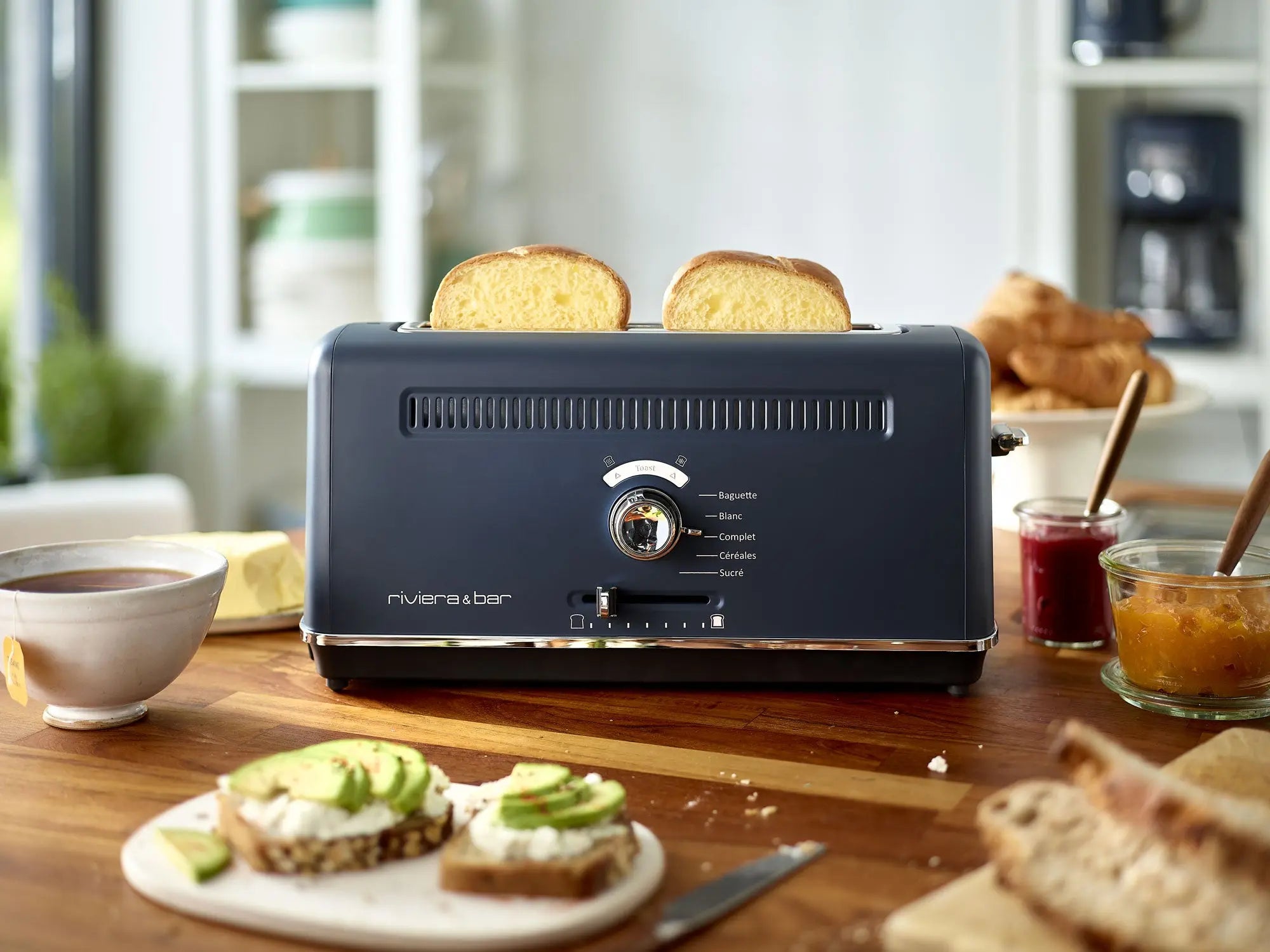 Un riviera&bar Grille-pain pour baguette avec fente extra large tient deux tranches de pain sur un comptoir de cuisine, à côté d'un café, d'un toast à l'avocat, de tranches de pain et de pots de confiture. Des étagères de cuisine apparaissent en arrière-plan.