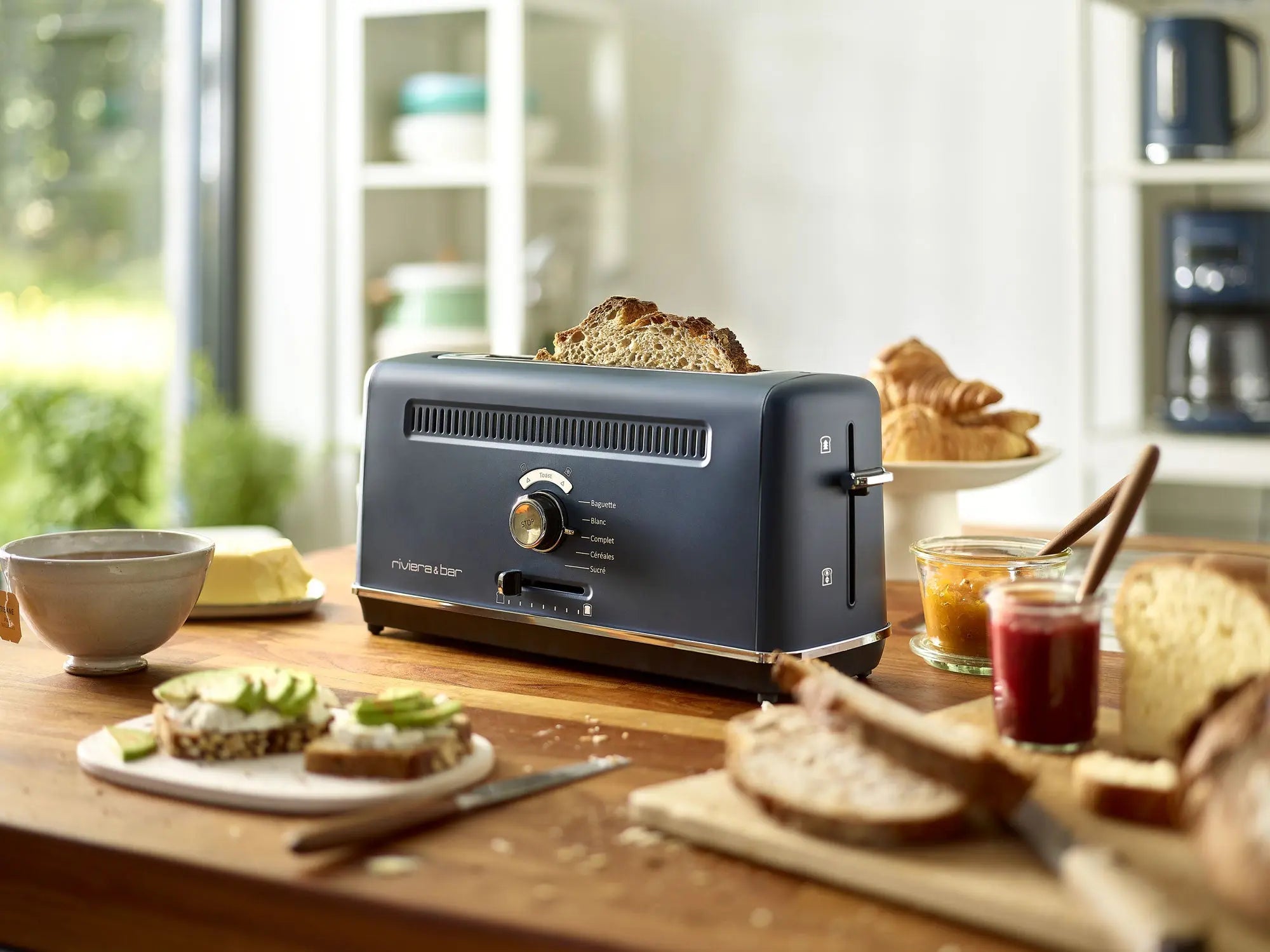 Le riviera&bar Grille-pain pour baguette avec fente extra large pops up pain sur un comptoir de cuisine en bois, entouré de pots de confiture, de beurre, d'un bol, et d'assiettes avec du pain tranché et de l'avocat. Des étagères et des appareils de cuisine apparaissent en arrière-plan.