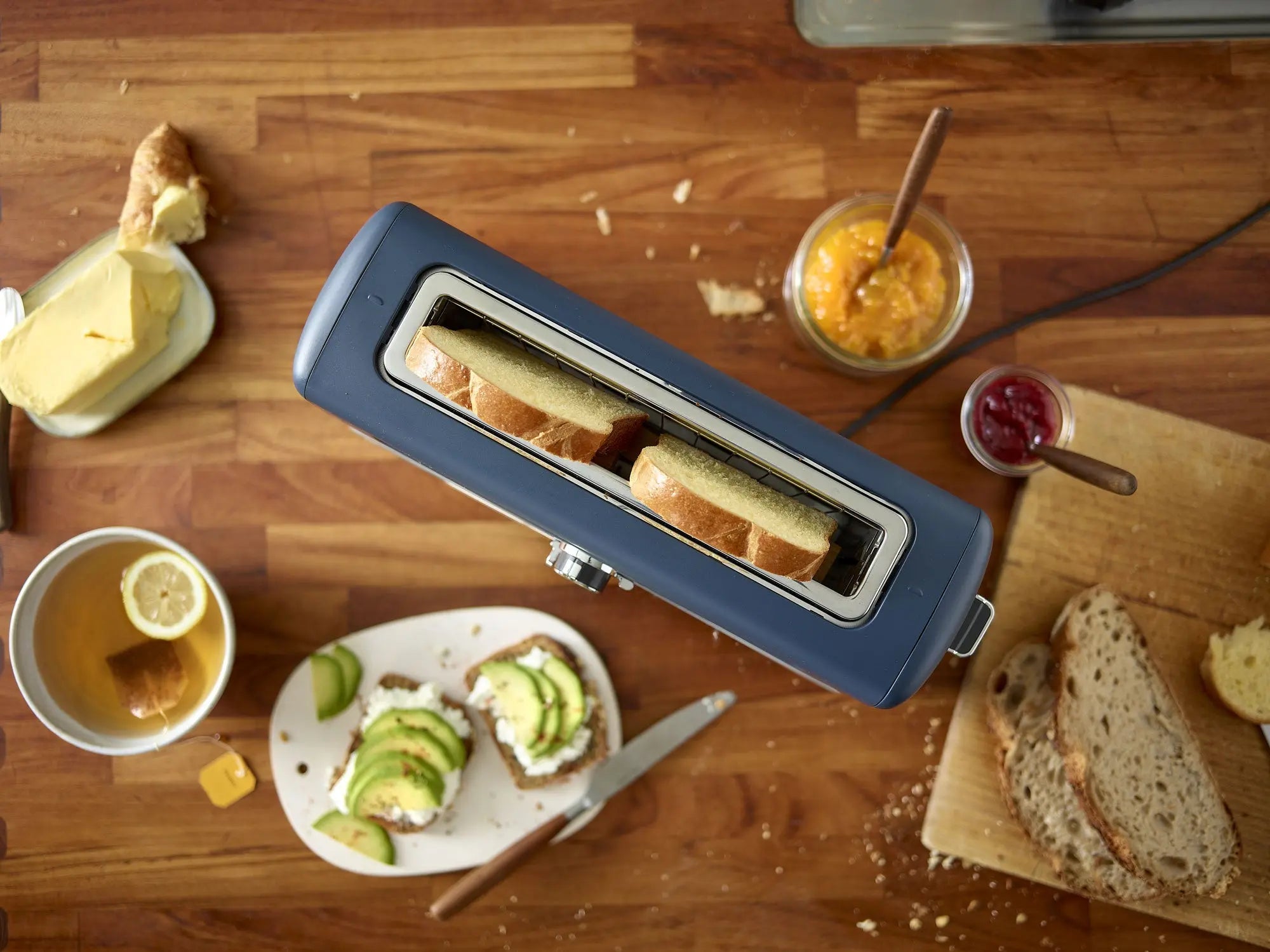 Vue de haut en bas du riviera&bar Grille-pain pour baguette avec fente extra large, entouré de beurre, de confiture, de pain tranché sur une planche à découper, d'une tasse de thé et d'un toast à l'avocat sur un comptoir en bois.
