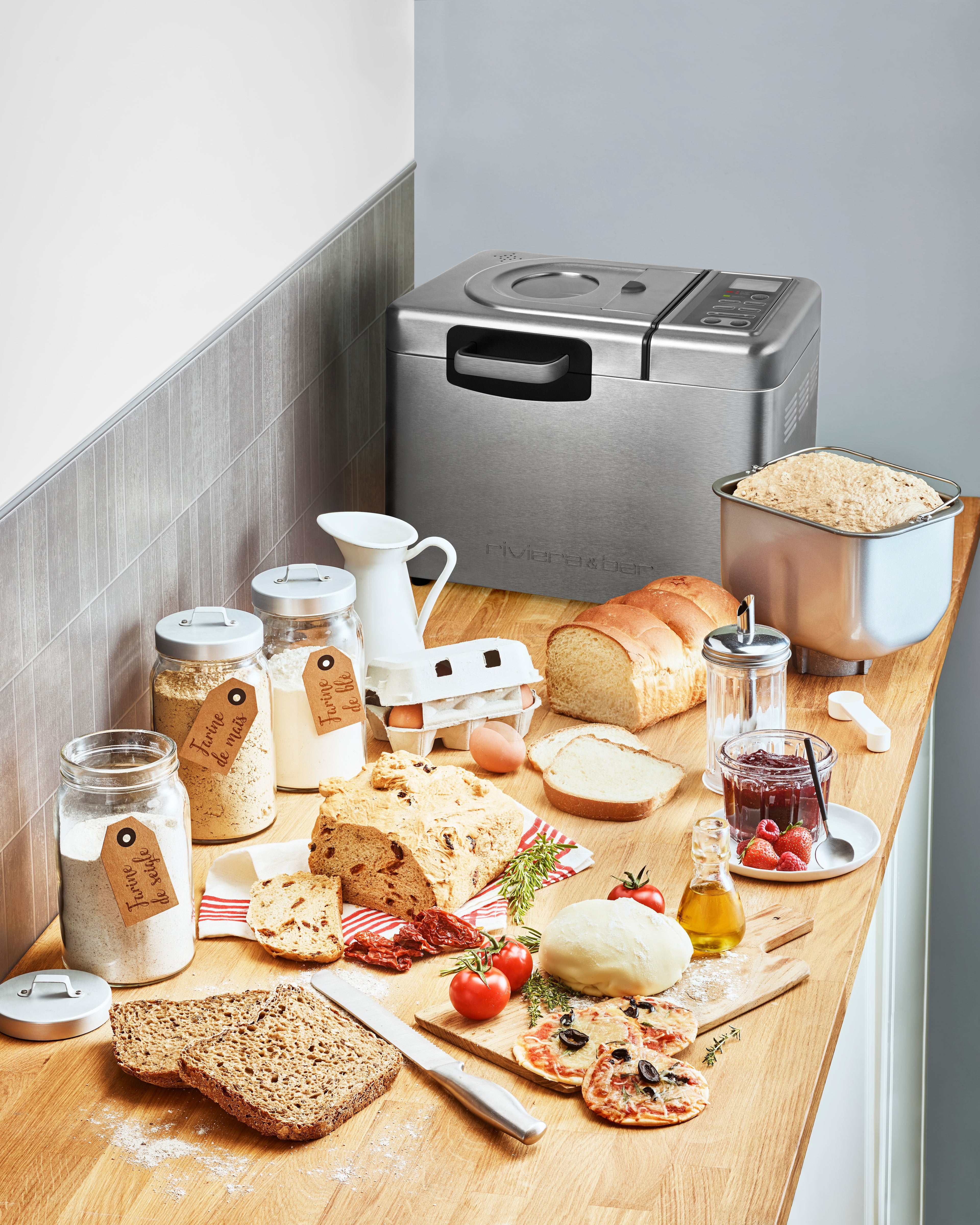 Sur un comptoir de cuisine lumineux et organisé se trouve la riviera&bar Machine à pain inox Sans gluten & Co - QMP 660, entourée de pain frais sans gluten, de bocaux de farine, d'œufs, de lait, de fromage, de tomates, d'herbes et de confitures - mettant en valeur les ingrédients et les produits de boulangerie.