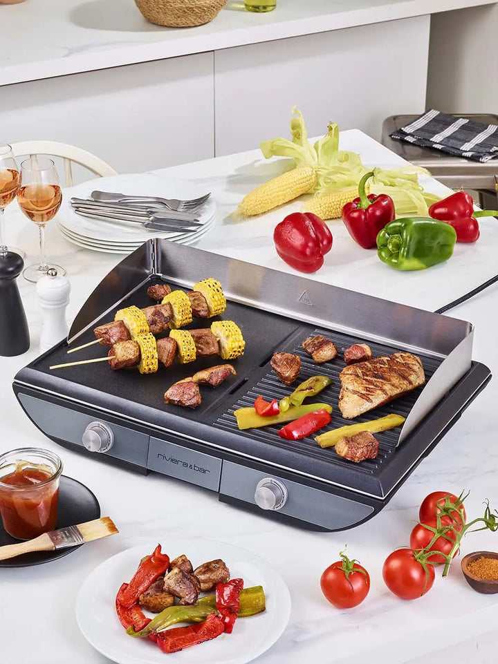 La Plancha 2 zones de cuisson riviera&bar dispose d'une surface de cuisson antiadhésive pour un nettoyage facile et est livrée avec des brochettes. Idéale pour griller du poulet, des légumes et du maïs dans une cuisine lumineuse. Verres à vin et assiettes sur le comptoir blanc.