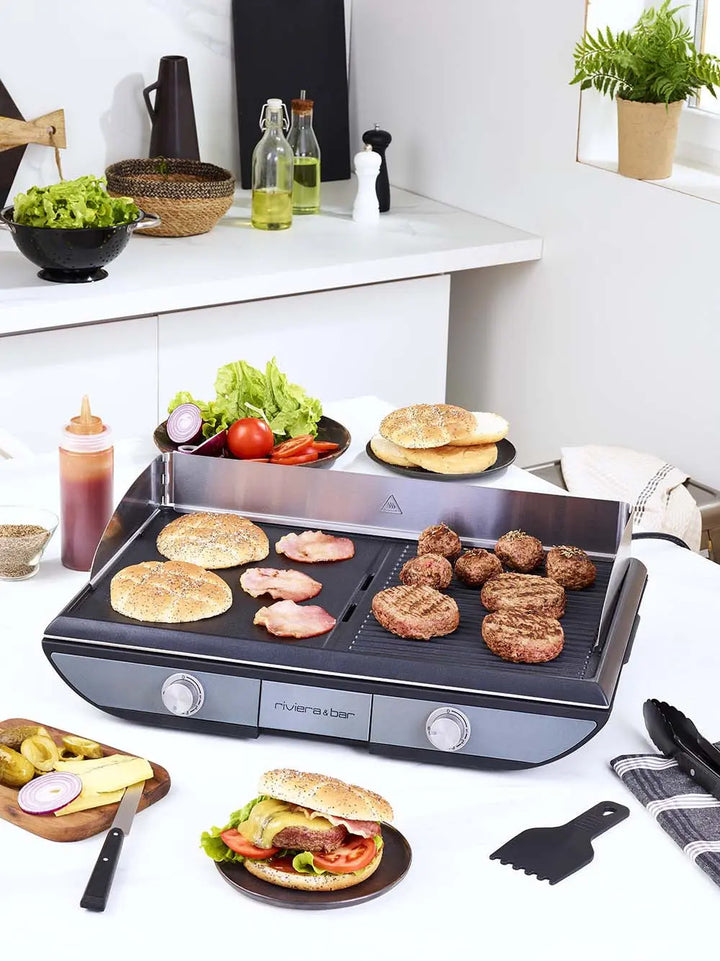 La Plancha 2 zones de cuisson riviera&bar, avec une surface antiadhésive, cuit facilement les galettes de hamburgers, les petits pains et le bacon. Des assiettes de légumes, des condiments et des hamburgers entourent le gril dans une cuisine moderne et lumineuse.