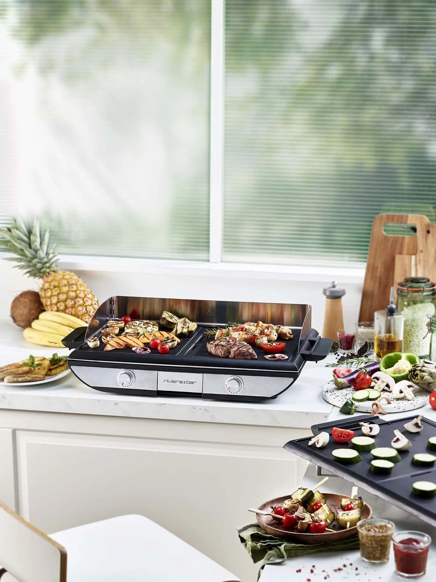 Une cuisine équipée de la riviera&bar Plancha électrique avec plaque amovible pour la cuisson des légumes et de la viande. Des fruits frais, des légumes en tranches et des brochettes entourent la plancha alors que la lumière du soleil filtre à travers les stores de la fenêtre pour une cuisson conviviale.