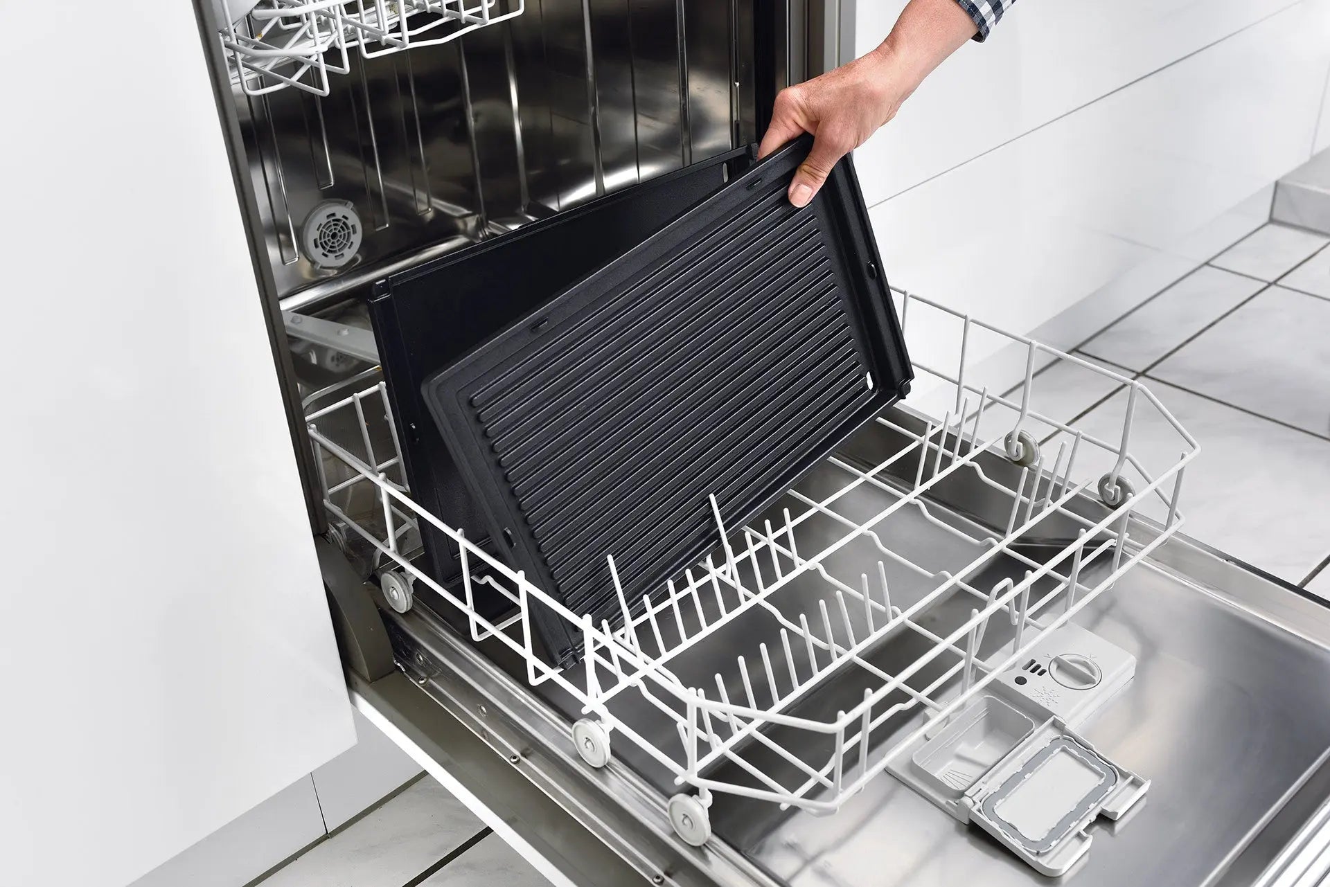 Une personne place une riviera&bar Plancha avec récupérateur de graisse sur la grille inférieure d'un lave-vaisselle ouvert dans une cuisine moderne avec des armoires blanches et un sol carrelé.