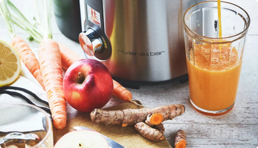 Gros plan sur des carottes fraîches, une pomme, des racines de curcuma et un citron à côté d'un verre de jus d'orange en train d'être versé, avec une centrifugeuse portant l'étiquette riviera & bar en arrière-plan.
