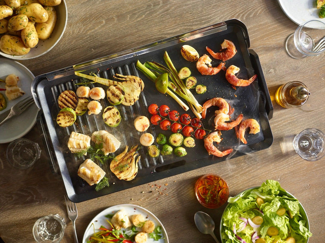 Un gril de table contenant des crevettes, des coquilles Saint-Jacques, des courgettes, des tomates, des oignons verts et du fenouil est entouré d'assiettes de salade, de pommes de terre, de verres à vin et de condiments sur une table en bois.