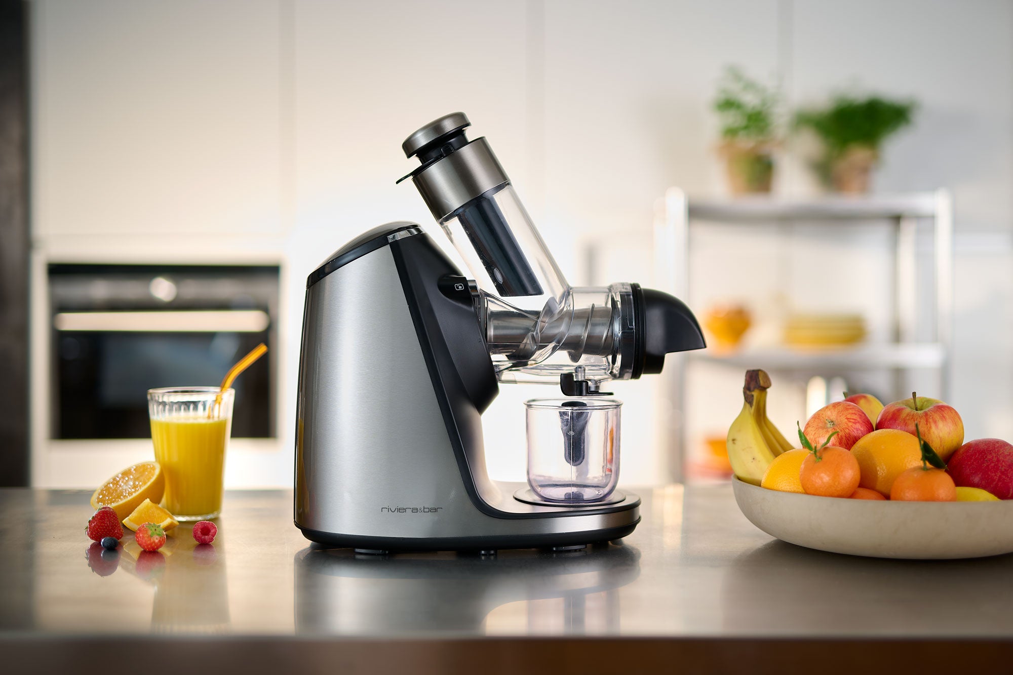 L'Extracteur de jus haute performance de riviera&bar est posé sur un comptoir de cuisine à côté d'un verre de jus d'orange avec une paille et des fruits frais, avec des fruits assortis et un arrière-plan de cuisine flou.