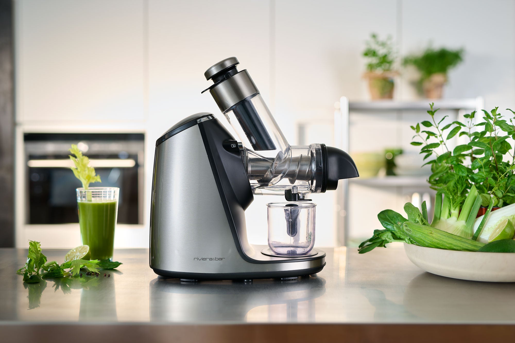 L'Extracteur de jus haute performance de riviera&bar est posé sur un comptoir de cuisine avec des légumes frais, des herbes et un verre de jus vert à proximité, mettant en valeur sa technologie d'extraction à froid contre des armoires modernes.