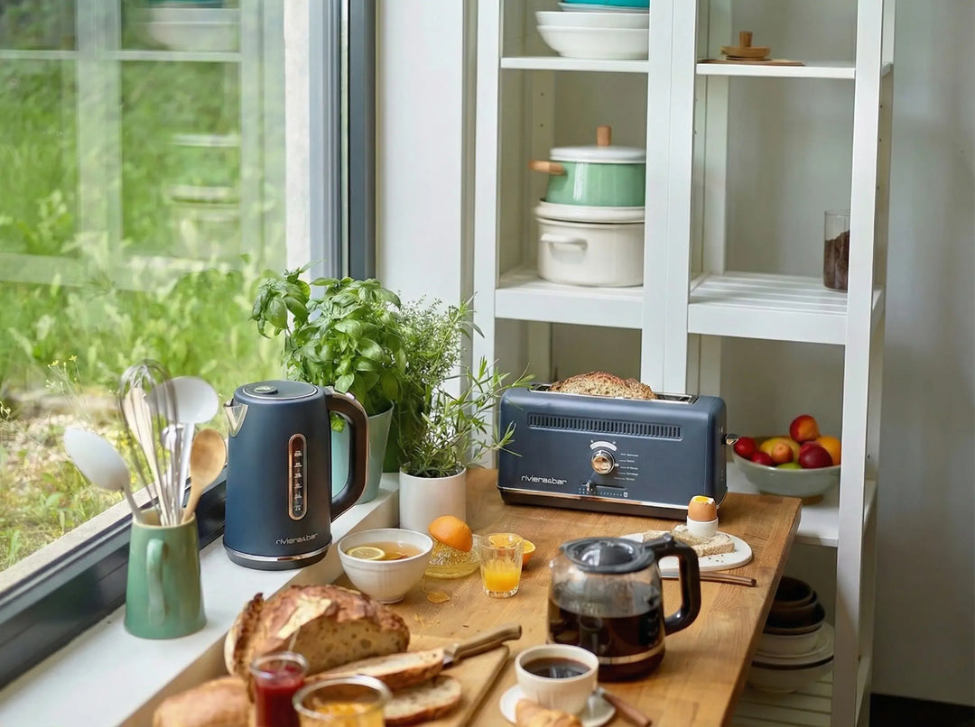 Une scène de cuisine confortable comprend une table en bois avec du pain, des pâtes à tartiner, une cafetière, riviera&bar Set bouilloire grille-douleur vintage, des tasses, et des aliments pour le petit déjeuner. La lumière du soleil pénètre à travers les herbes en pot et les étagères bordées de vaisselle en arrière-plan.
