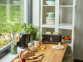Un comptoir de cuisine lumineux près d'une fenêtre présente la riviera&bar Set bouilloire grille-pain noir avec du pain, des herbes, des assiettes de fruits, des bocaux, des ustensiles et des jus de fruits, créant ainsi le parfait petit-déjeuner. Les étagères situées à l'arrière contiennent des plats et des bols.