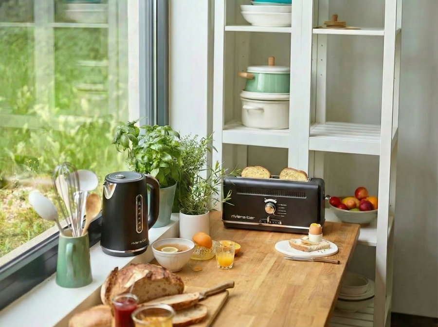 Un comptoir de cuisine lumineux près d'une fenêtre présente la riviera&bar Set bouilloire grille-pain noir avec du pain, des herbes, des assiettes de fruits, des bocaux, des ustensiles et des jus de fruits, créant ainsi le parfait petit-déjeuner. Les étagères situées à l'arrière contiennent des plats et des bols.