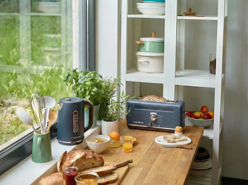 Un plan de travail en bois, sur lequel sont disposés du pain, de la confiture, du jus de fruits, des œufs, un ensemble « Duo Bouilloire & Grille-Pain » de couleur gris-bleu de la marque Riviera&Bar, des herbes aromatiques fraîches et des ustensiles de cuisine, se trouve près d'une fenêtre donnant sur le jardin ; à proximité, des étagères blanches présentent de la vaisselle et des tomates — l'idéal pour préparer votre petit-déjeuner.