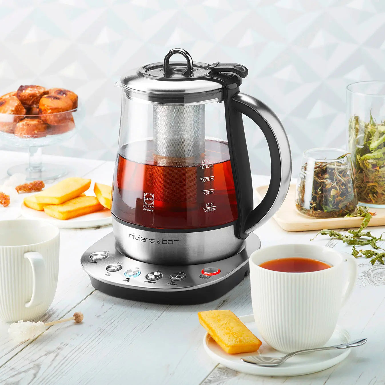 Théière électrique en verre et acier inoxydable, accompagnée de tasses, de gâteaux et d'ingrédients pour le thé.