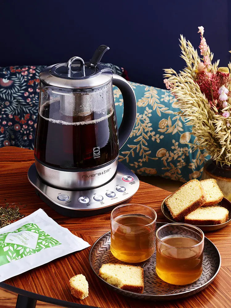 Une Théière Jaïpur Zen (1,2 L) de riviera&bar remplie de thé noir est posée sur une table en bois à côté de deux tasses en verre de thé infusé, de tranches de gâteau, d'un sachet de thé et d'un bouquet de fleurs séchées en arrière-plan.
