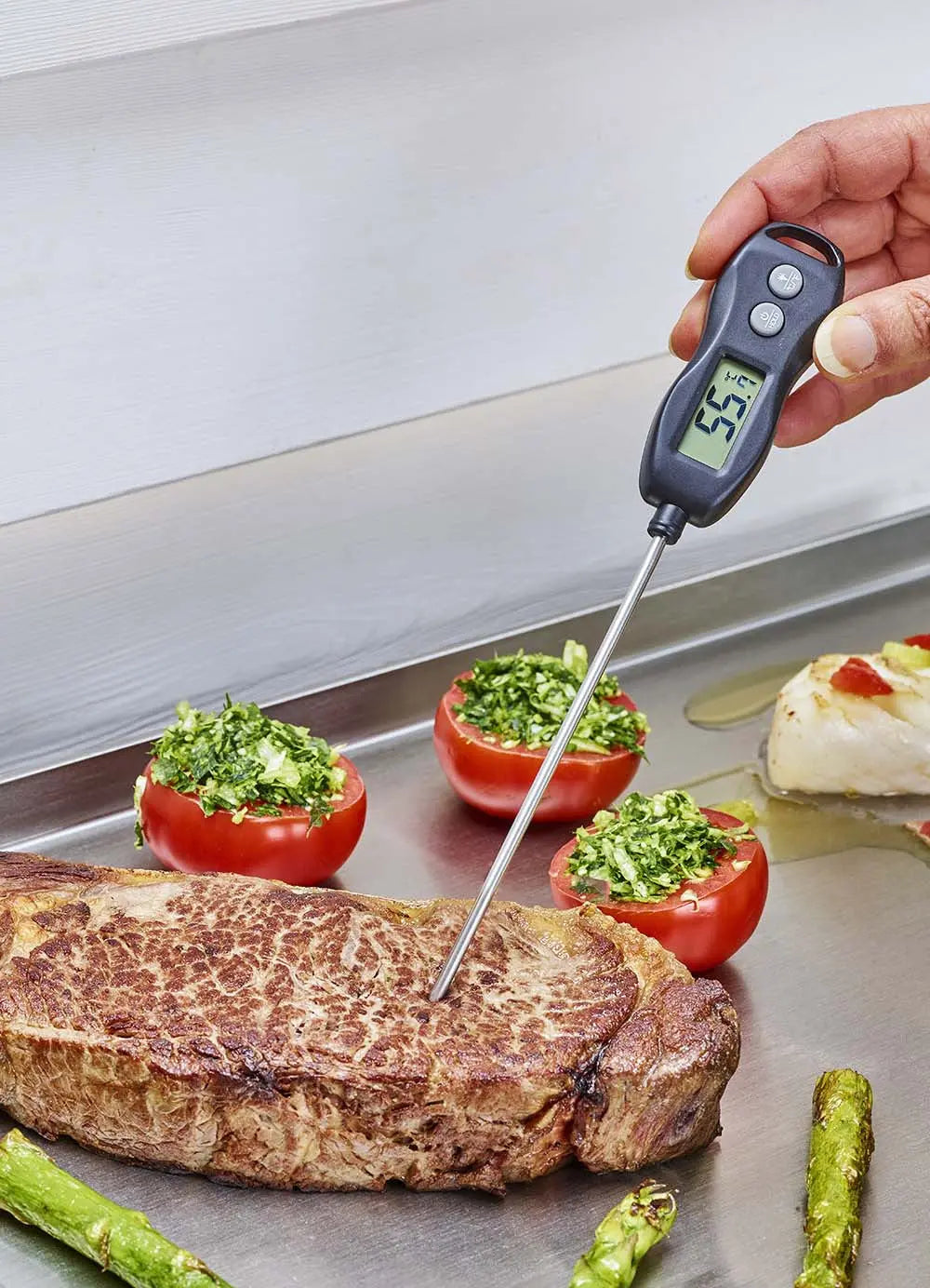 Thermomètre de cuisson mesurant la température d'un steak grillé, tomates farcies et asperges à proximité.
