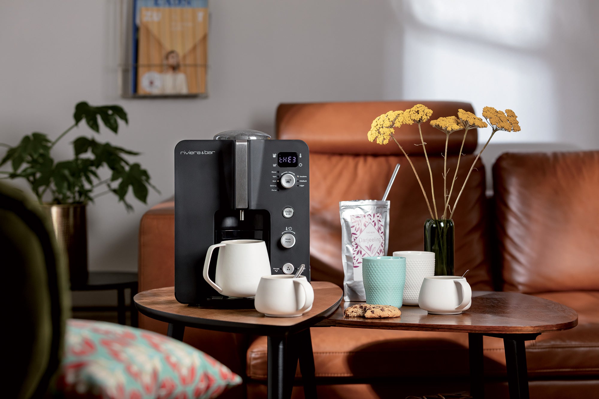 Une cafetière est posée sur une table en bois avec des tasses, un gobelet, des biscuits et un vase de fleurs jaunes dans un salon confortable avec un canapé en cuir marron et une plante en arrière-plan.